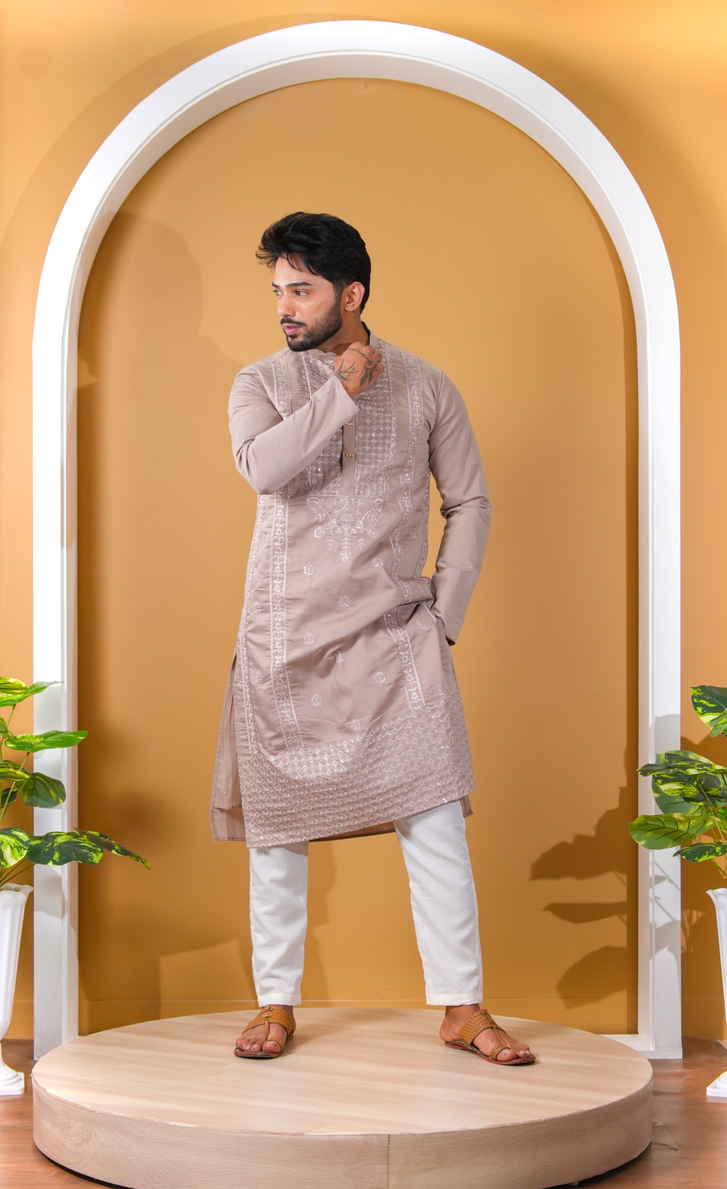 Man wearing a beige kurta with white pants standing in front of a yellow wall with an arch.