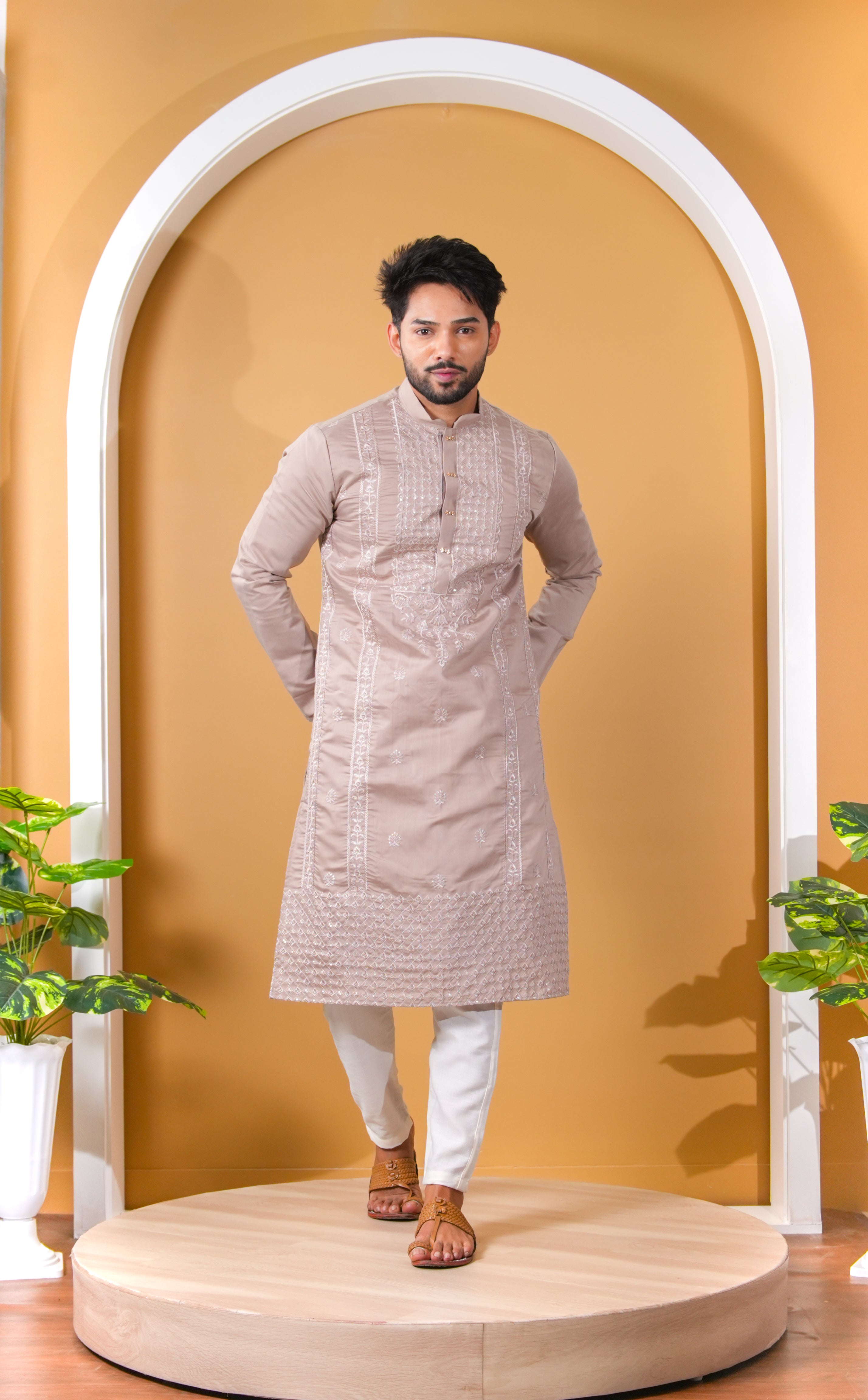 Man wearing a beige kurta with white pants standing in front of a yellow wall with an arch.