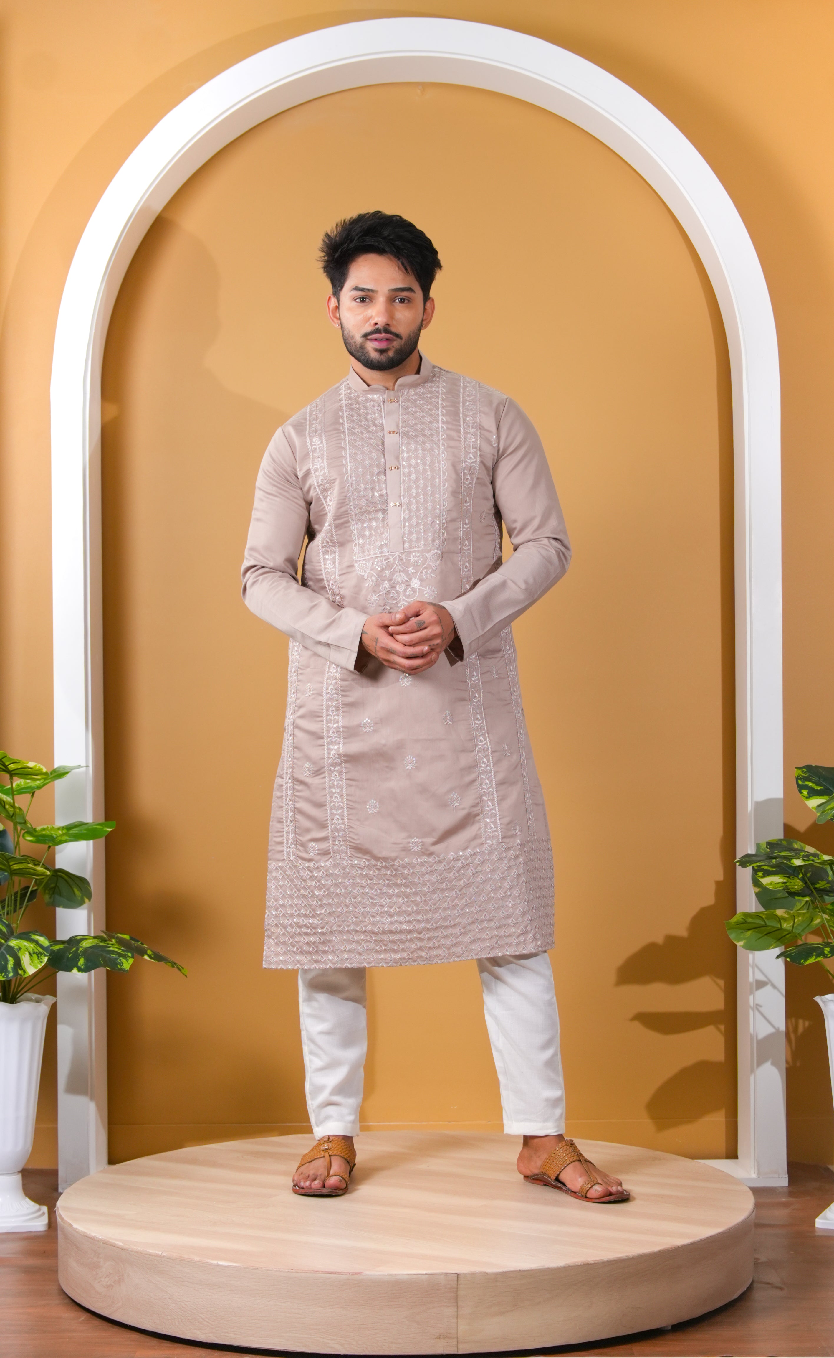 Man wearing a beige kurta with white pants standing in front of a white archway on a yellow background.