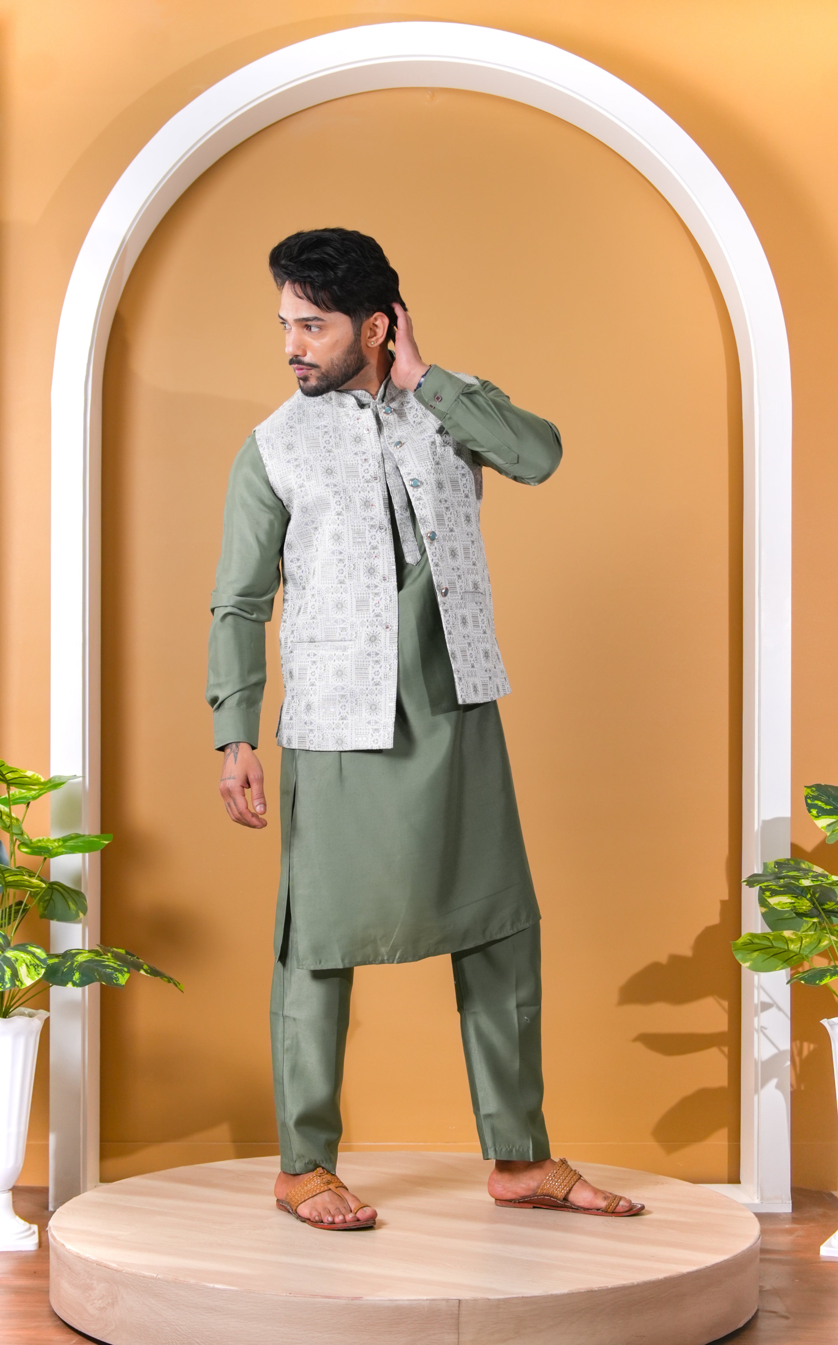 Man wearing a green traditional outfit with a white vest against a beige wall.
