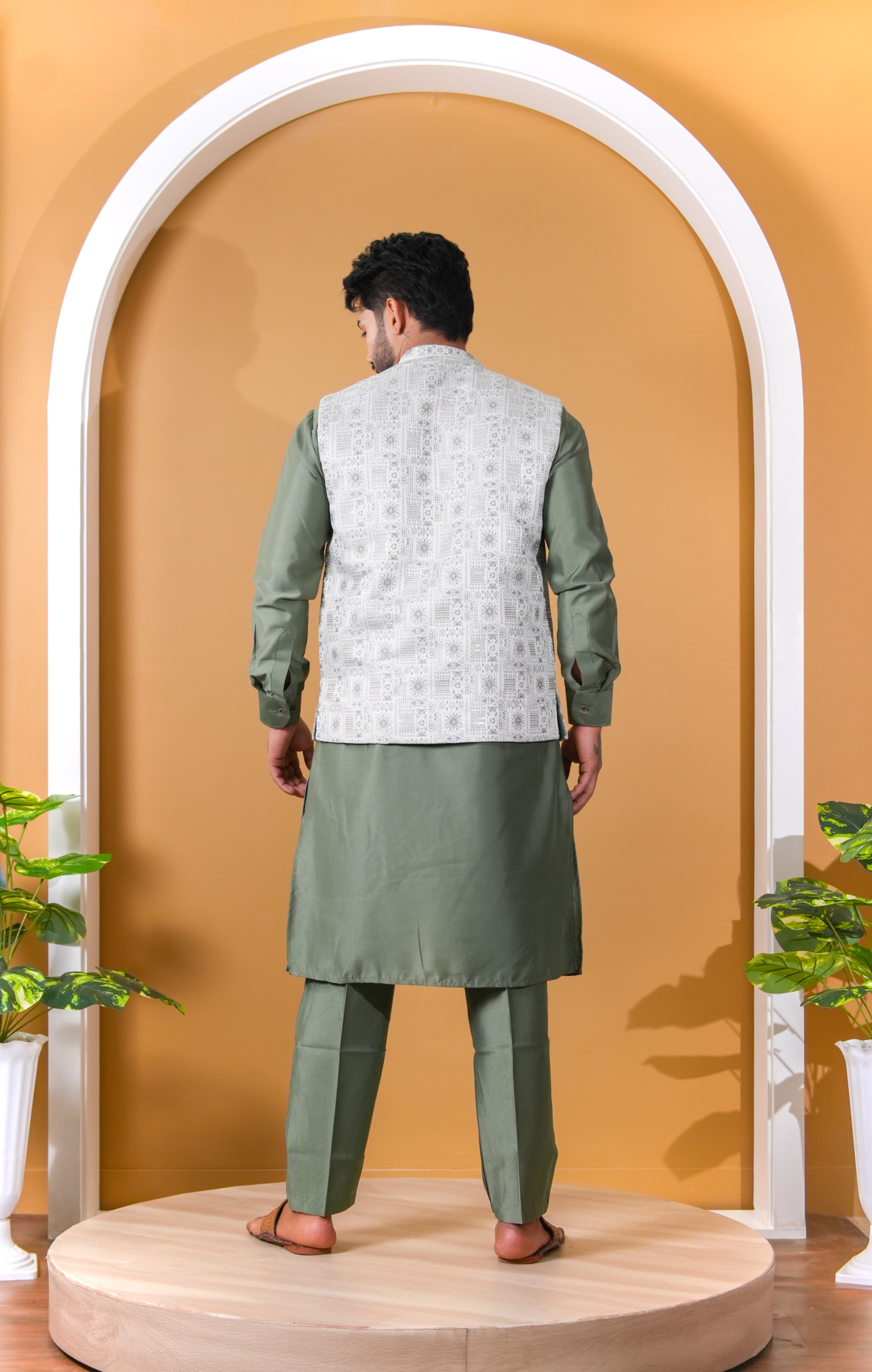 Man wearing a green kurta with a white embroidered jacket against an orange wall.