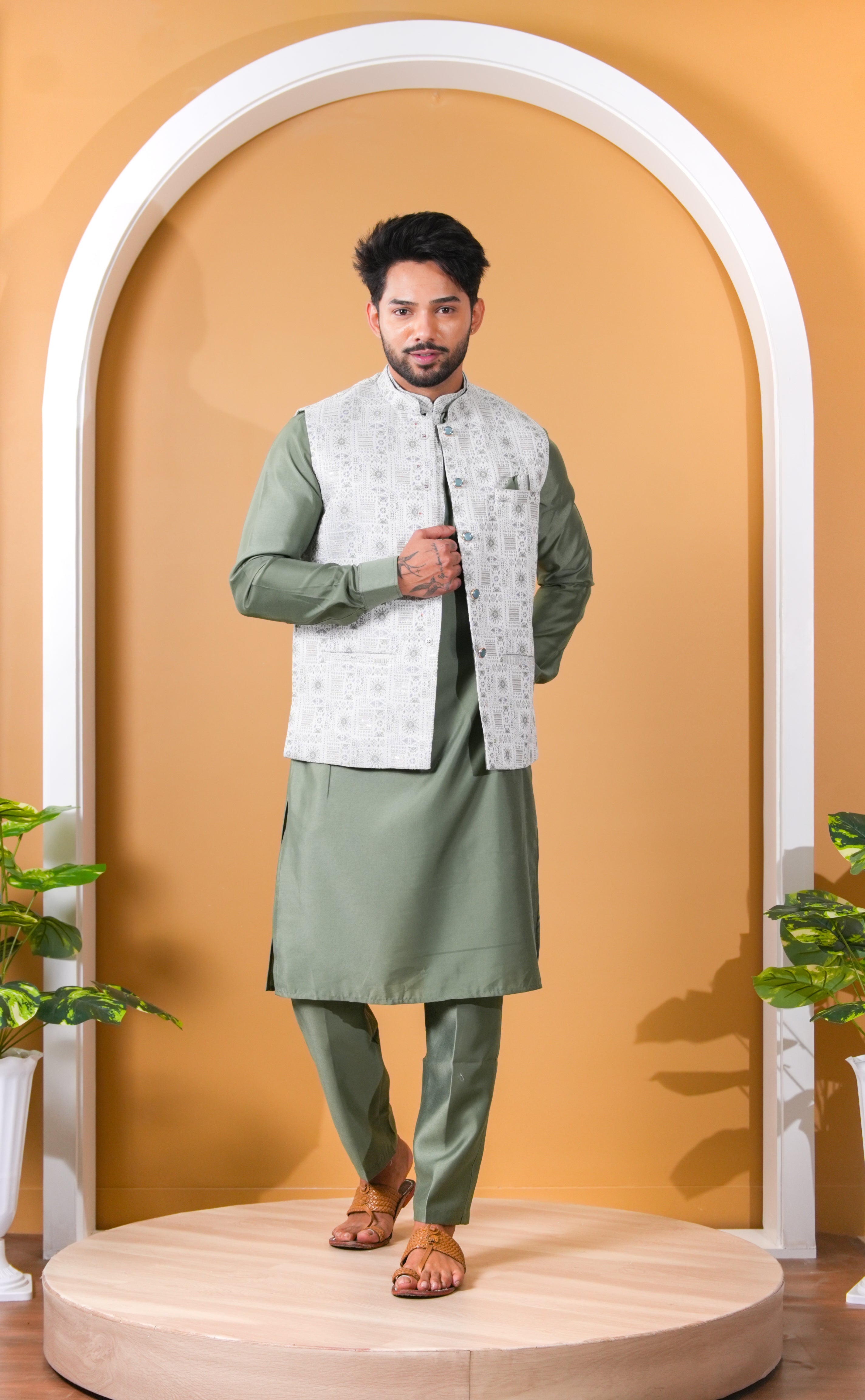 Man wearing a green kurta with a white embroidered jacket against a beige wall.