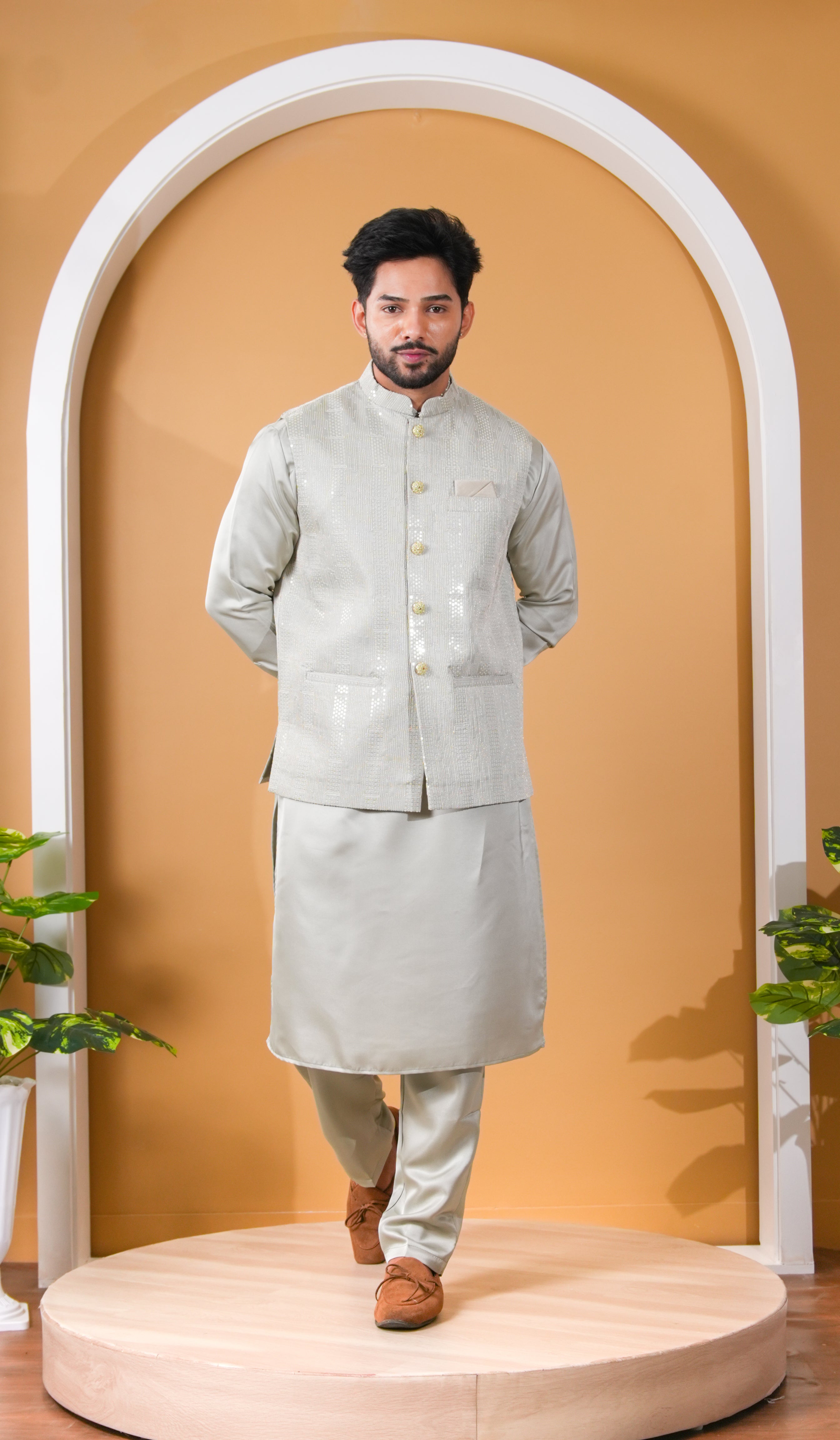 Man wearing a light gray traditional outfit standing in front of a white archway with a beige wall.
