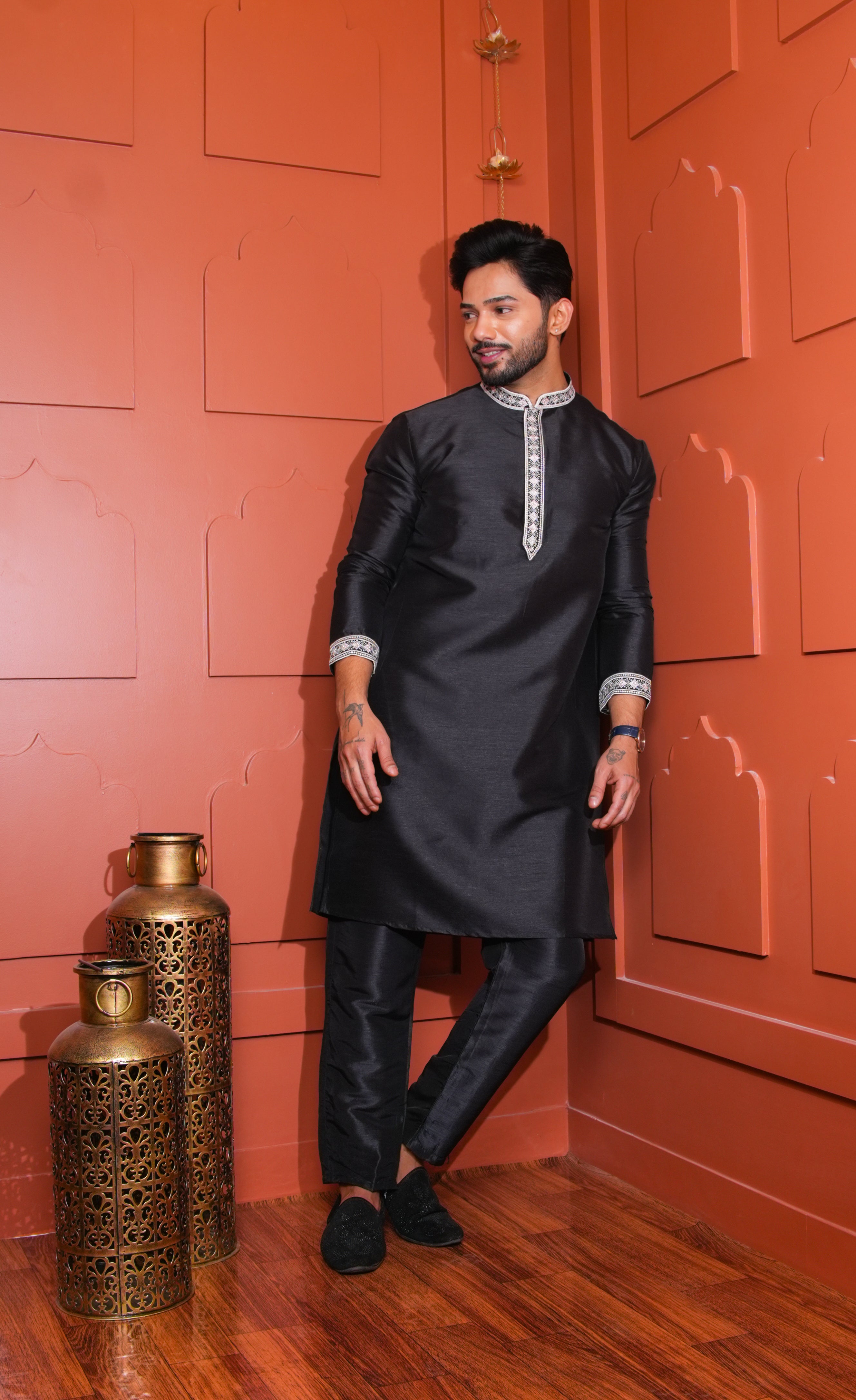 Man in a black kurta standing against an orange wall with decorative elements.