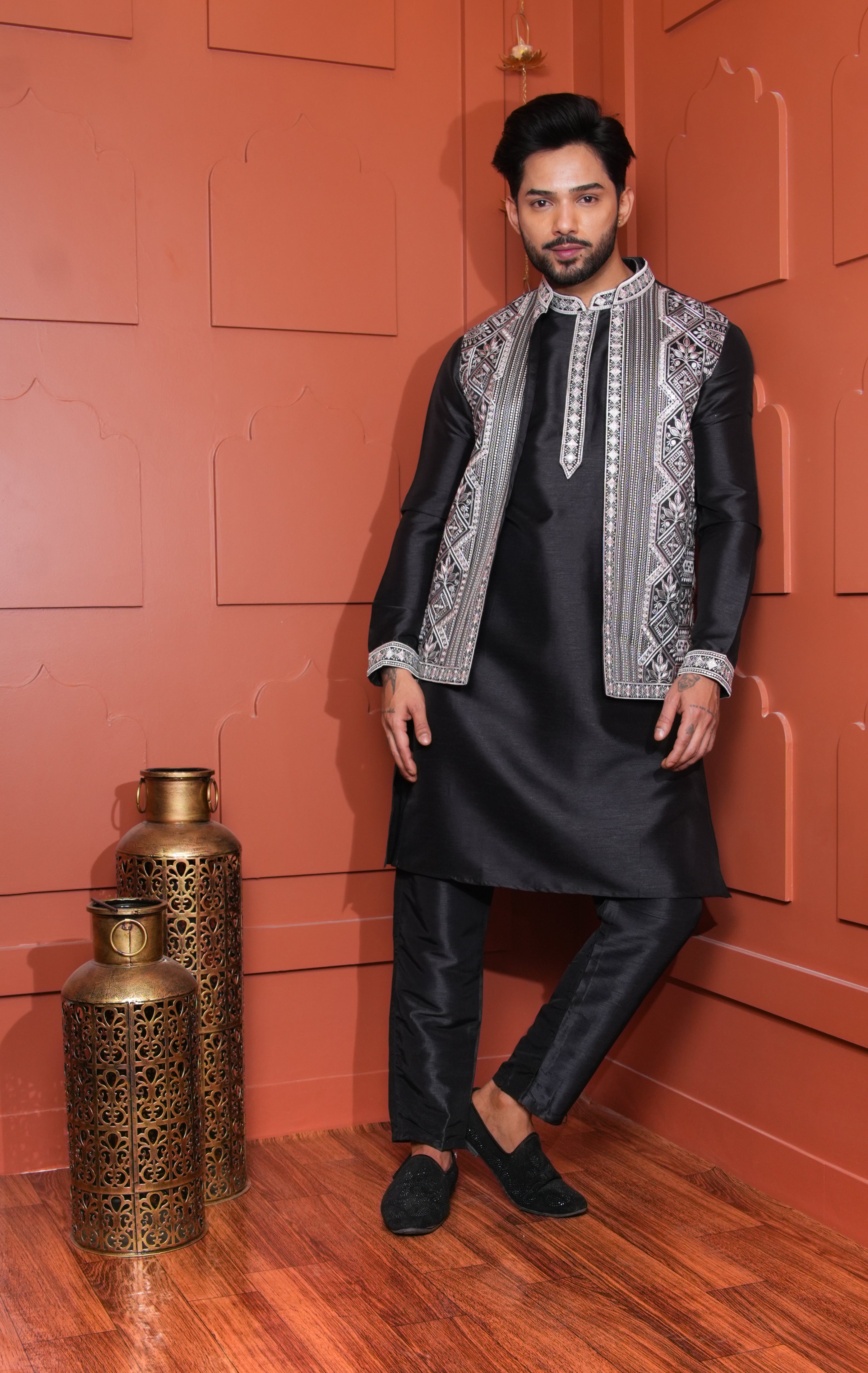 Man in traditional black outfit with silver embroidery standing against a red wall with decorative vases.