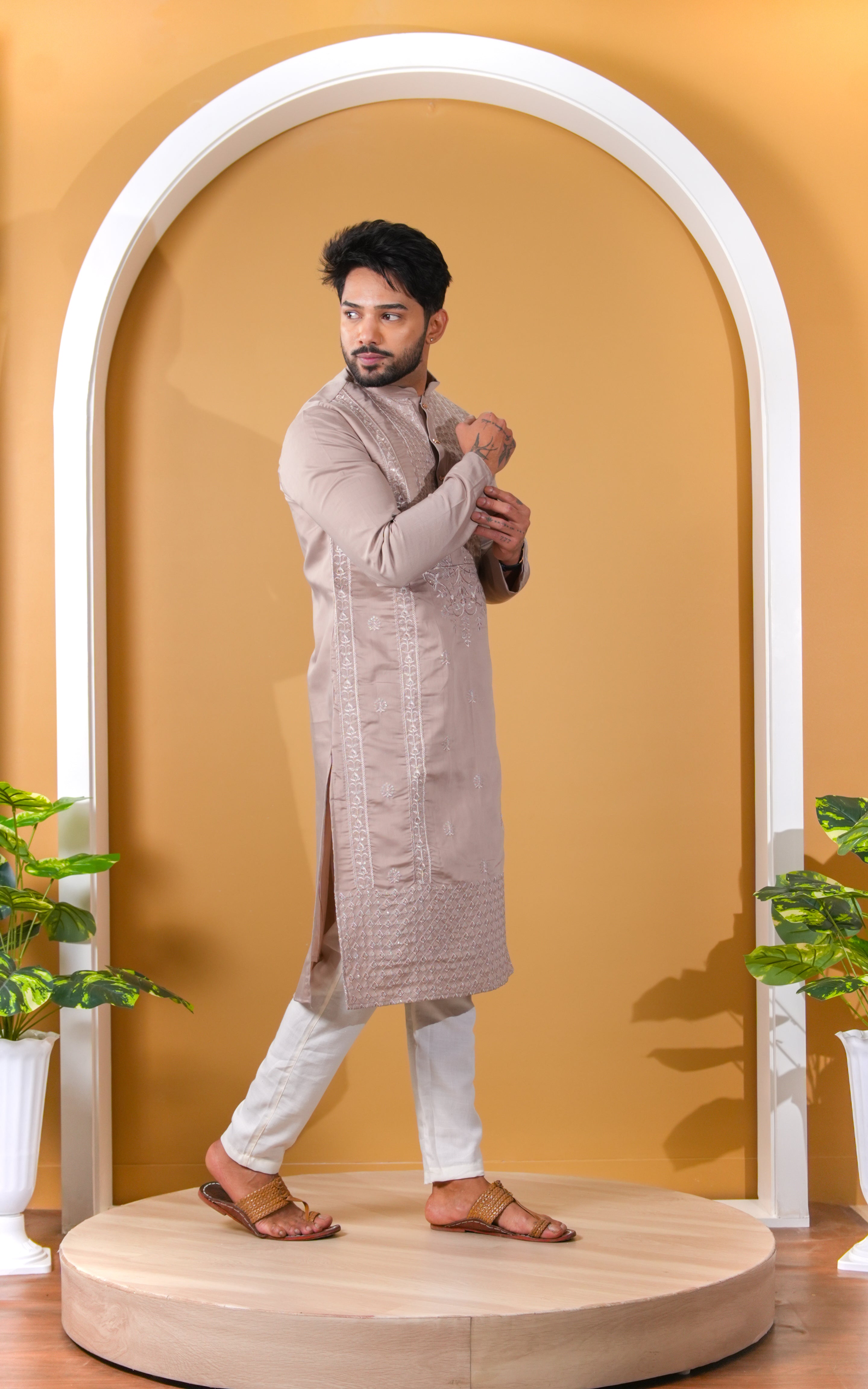 Man wearing a traditional kurta standing in front of an archway with a yellow wall and plants on either side.
