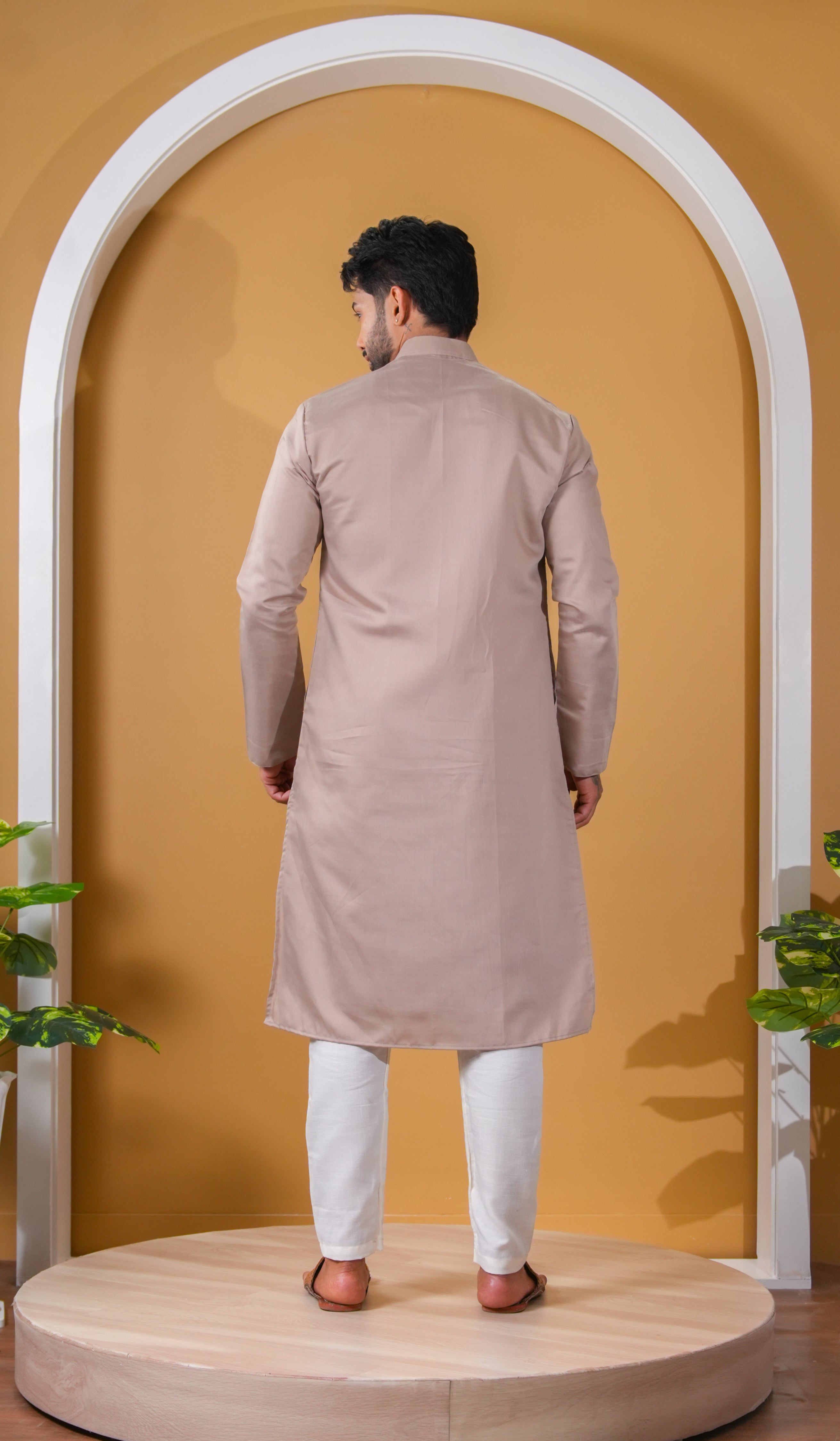 Man wearing a beige kurta with white pants standing in front of a yellow wall with an arch.