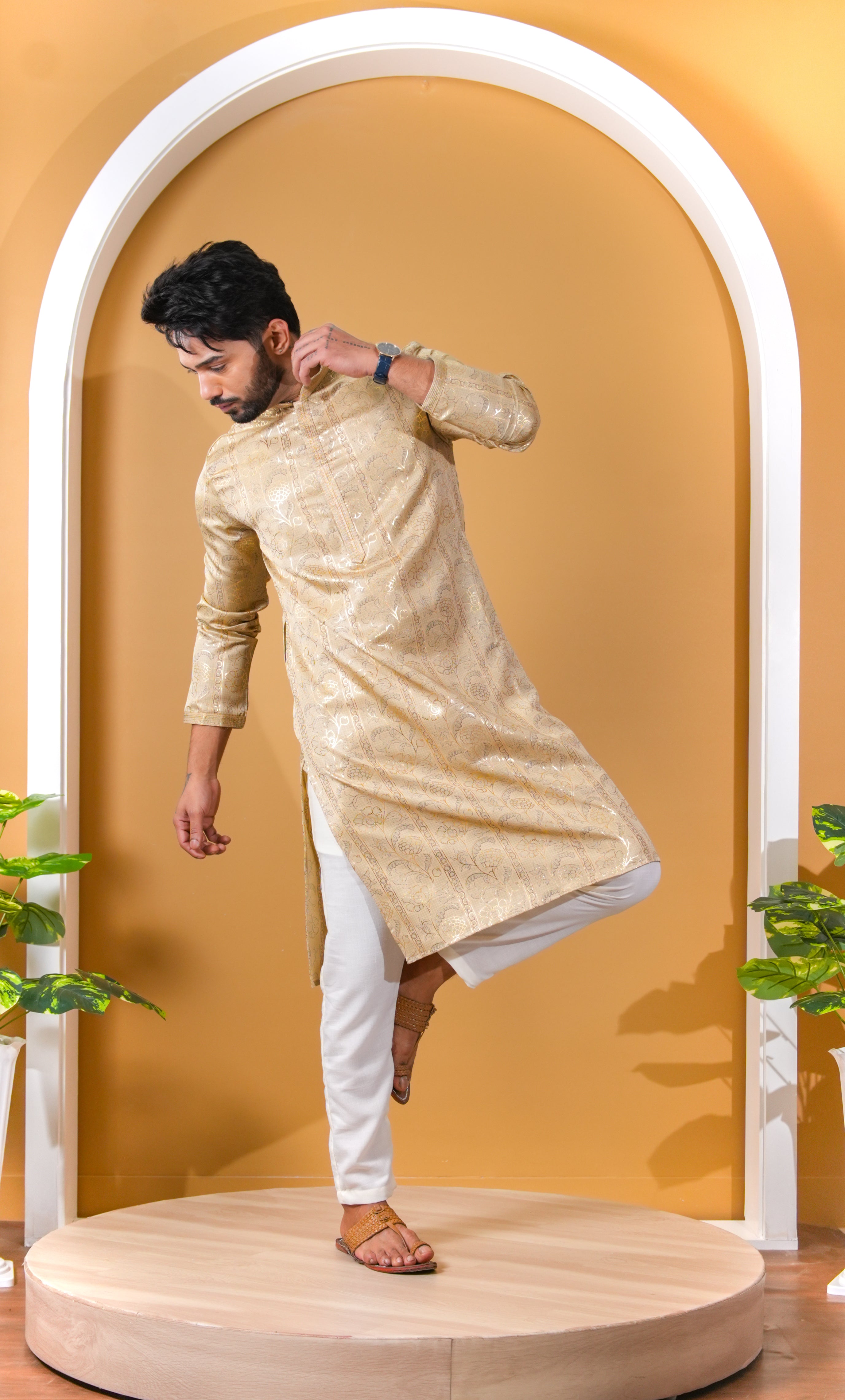 Man wearing a beige kurta with white pants standing on a wooden platform against a yellow wall.