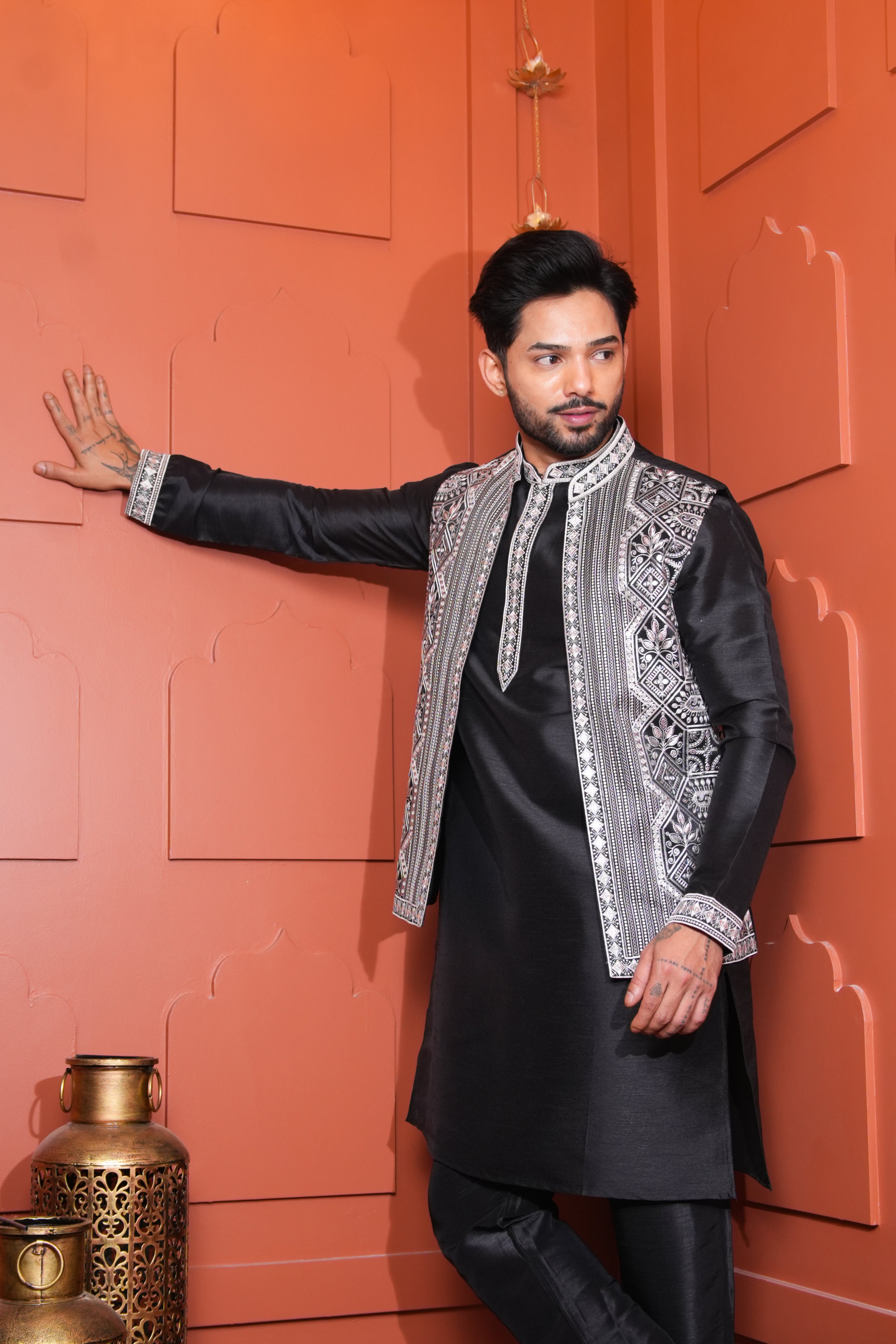 Man wearing a black and silver embroidered traditional outfit against an orange wall.